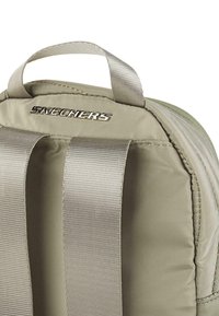 Mochila Skechers color caqui con un acabado de tela suave, logotipo metálico y correas anchas y texturizadas. Cuenta con un asa superior para mayor comodidad.