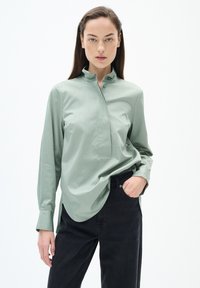 Hellgrühes Hemd mit Knopfleiste, hohem Kragen, langen Ärmeln und einer Vorderseite mit Knopfleiste. Getragen mit dunklen Jeans. Glatte Textur, lässiges Design.