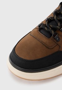 Bruin suede en zwart stoffen sneaker met een textuur, contrasterende rubberen zool en zwarte en gele veters.