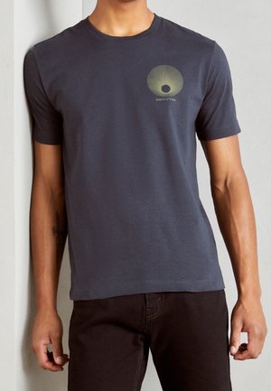 Mann trägt ein dunkelgraues kurzärmeliges T-Shirt mit einem kleinen gelben runden Design und Text auf der oberen linken Brust.