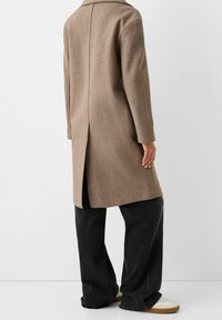 Cappotto di lana marrone con taglio dritto, colletto a lobo e spacco sul retro, abbinato a jeans neri a gamba larga e sneakers chiare.