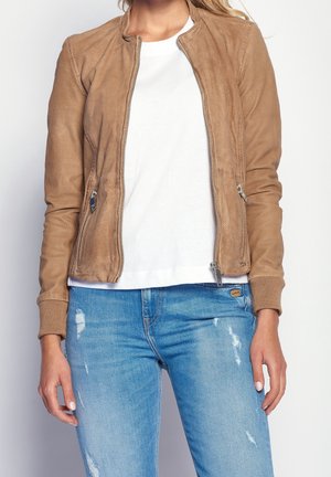 Woman wearing a tan perforated zip-up jacket over a white shirt with distressed blue jeans, standing against a plain background.