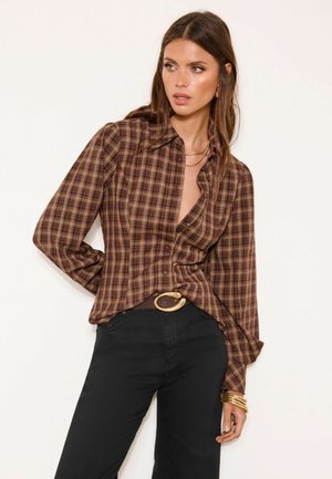 Frau trägt ein braunes kariertes Hemd mit Knöpfen, das in hoch taillierte schwarze Hosen gesteckt ist, dazu ein großer Gürtel mit goldener Schnalle und goldene Armbänder.