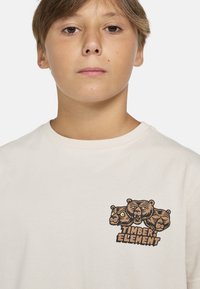 Beige t-paita, jossa on graafinen printti kolmesta karhun päästä ja "TIMBER ELEMENT" -teksti ruskeana, klassisella pyöreällä kaula-aukolla ja pehmeällä tekstuurilla.