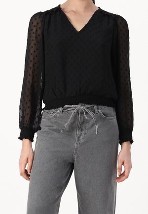 Femme portant un chemisier noir transparent à pois avec manches longues et poignets volantés, ainsi qu'un jean gris taille haute avec ceinture à cordon.