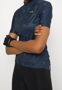 Cyklistický dres s krátkým rukávem v tmavě modré barvě s bílými křížovými vzory, předním zipem, jemným logem a přiléhavým střihem.