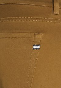 Bruna byxor med en slät textur, med kontrasterande vita och svarta ränder på överkanten av vänster ficka och synliga sömnadsdetaljer.