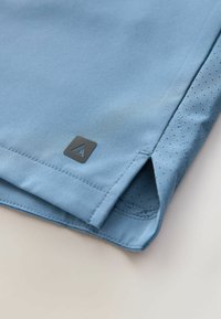 Ourlets de shorts de sport bleu clair avec fente latérale et doublure intérieure perforée, ornés d'un petit patch logo triangulaire noir.
