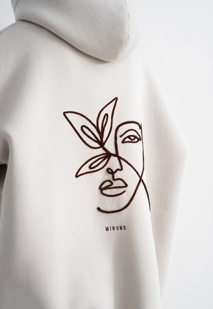 Hoodie in lichtbeige met een bruine geborduurde abstracte gezicht en bladeren ontwerp; zachte stof met een ontspannen pasvorm.