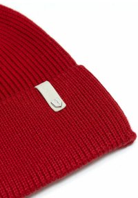Czerwona dziergana czapka z prążkowaną fakturą, z białą prostokątną metką na mankiecie z logo.