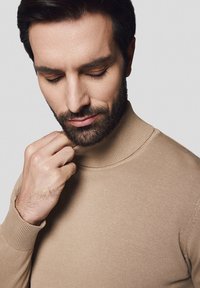 Beżowy sweter z golfem, wykonany z prążkowanego materiału, o dopasowanym kroju i wysokim kołnierzu, noszony przez osobę z krótkimi włosami i zarostem.