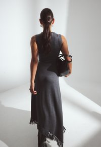 Naine pikkade tumedate juustega, kühveldatud kuklaga, seljas õlgadeta, asümmeetriline tumehall kleit ja käes must kott.