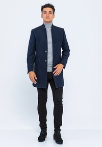 Donker marineblauwe overjas met een getailleerde pasvorm, voorzien van een hoge kraag, twee knopen en zijzakken, gedragen over een lichtgrijze turtleneck.