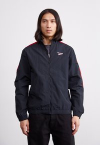 Chaqueta cortaviento negra con acentos rojos, cierre de cremallera y puños elásticos. Textura suave, con un logo en el lado izquierdo del pecho.