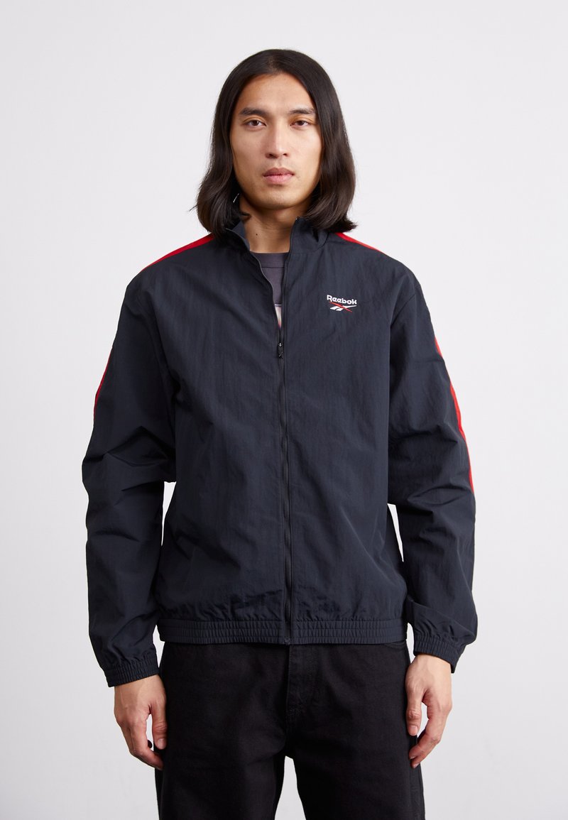 Chaqueta cortaviento negra con acentos rojos, cierre de cremallera y puños elásticos. Textura suave, con un logo en el lado izquierdo del pecho.