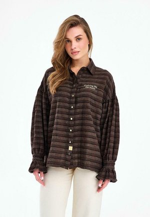 Kobieta ubrana w luźną, ciemnobrązową, w paski koszulę na guziki z falbaniastymi mankietami oraz beżowe spodnie, stojąca na tle białej ściany.