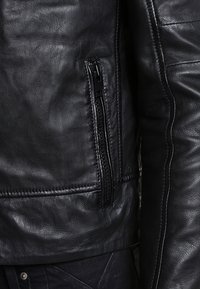 Schwarze Lederjacke mit strukturierter Oberfläche, Reißverschlusstasche und taillierter Passform mit sichtbaren Nähten entlang der Seiten.