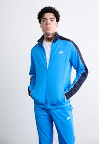 Βασιλικό μπλε φόρμα Nike με ζακέτα με φερμουάρ και παντελόνια με ελαστική μέση. Διαθέτει ναυτικές λεπτομέρειες, λευκό λογότυπο και πλαϊνές τσέπες. Λείος υφάσματος.