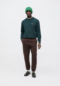 Dark green sweatshirt with a small logo, brown tapered sweatpants featuring side text, green knit beanie, and black loafers with white socks.