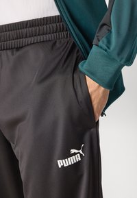 Svarte treningbukser med elastisk liv, sidelomme og hvit "PUMA"-logo på nederste del av beinet. Glatt, lett stoff.