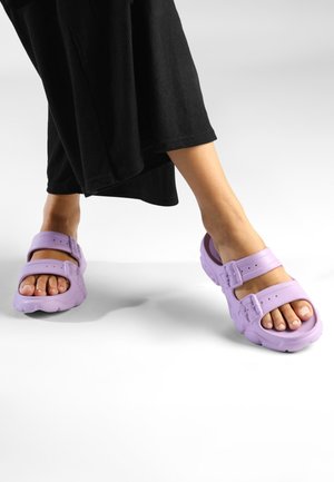Purple rubber sandals with two adjustable straps, thick soles, and a textured surface. Worn with a black ribbed garment.