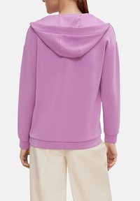 Sudadera púrpura con capucha, puños y dobladillo acanalados. Tela suave y lisa con un corte relajado, combinada con pantalones de color claro.