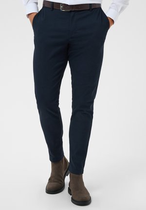 Bărbat purtând pantaloni conici de culoare bleumarin închis, curea maro și ghete maro din piele întoarsă, stând cu mâinile în buzunare.