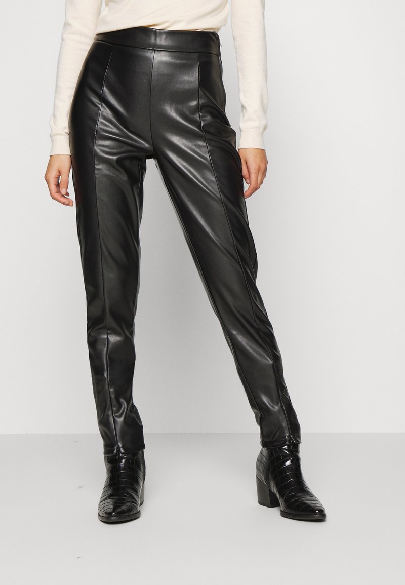 Nainen, jolla on päällään mustat tekonahkahuoneet ja mustat nilkkurit, seisoo tasaisella vaalealla taustalla.