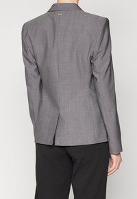 Blazer gris à coupe ajustée, fente unique au dos et motif texturé subtil. Comprend une étiquette de marque au niveau du col.