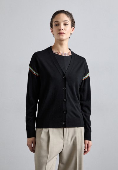 Cardigan noir avec un décolleté en V, fermeture par boutons et accents multicolores rayés sur les manches. Porté sur un top noir et un pantalon beige.