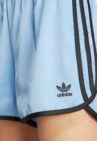 Ljusblå idrottsshorts med svarta detaljer, med Adidas-logotypen i svart på nedre vänstra sidan. Tillverkad av ett mjukt, glänsande material.