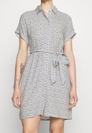 Robe à manches courtes avec un motif complexe noir et blanc, un col et une ceinture en tissu nouée à la taille, portée par une personne avec des tatouages sur les bras.