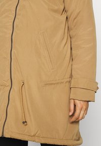 Parka beige avec fermeture éclair frontale, poche poitrine inclinée, taille réglable par cordon et sangle de poignet sur la manche droite.