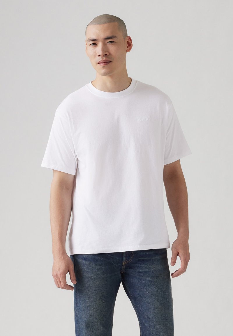 Hvit bomulls T-skjorte med korte ermer og rund hals, som har en subtil, brodert logo på brystet. Brukt sammen med blå jeans.