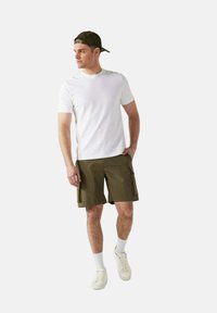 Witte t-shirt met korte mouwen, gecombineerd met olijfgroene cargoshorts, witte crew-sokken en witte sneakers, tegen een neutrale achtergrond.