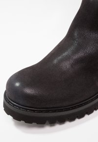 Gros plan sur une botte en cuir noir avec un bout arrondi et une semelle épaisse en caoutchouc à relief sur fond blanc.