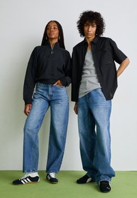 Svart quarter-zip sweatshirt, lys grå tank top og mørk skjorte kombinert med vide støvler i blå jeans. Begge har på seg joggesko i kontrastfarger.