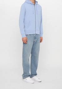 Ljusblå zip-up huvtröja med huva, framfickor och logobroderi; bärs med ljusblå raka jeans och vita sneakers.