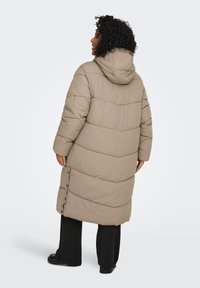 Lång, vadderad kappa i en ljusbeige färg med huva. Har ett quiltat mönster, sidoknappar och en avslappnad passform.