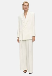 Blazer bianco oversize con rever a punta e tasche frontali, abbinato a pantaloni a gamba larga. Tessuto dalla texture liscia, design minimalista.