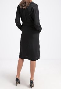 Černý trench coat s páskem v pase, dlouhými rukávy a délkou po kolena. Vyznačuje se hladkým texturem a klasickým límcem.