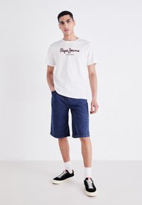 Hvit t-skjorte med kort erm og "Pepe Jeans London" logo, sammen med knelengde blå denimshorts og svarte sneakers med hvite såler.
