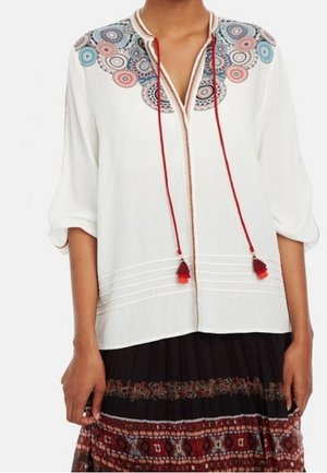 Mujer con una blusa blanca bordada con círculos de colores en los hombros y lazos con borlas rojas, combinada con una falda estampada.