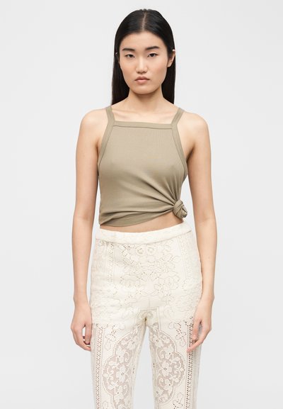 Γυναίκα με μακριά μαύρα μαλλιά που φοράει ένα μπεζ αμάνικο τοπ δεμένο στη μέση και παντελόνι με ψηλή μέση σε λευκό χρώμα με δαντέλα, πάνω σε λευκό φόντο.