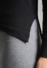 Maglietta nera a maniche lunghe con dettagli all'orlo curvo, sovrapposta a un tessuto grigio, caratterizzata da una texture liscia e cuciture visibili lungo le cuciture.