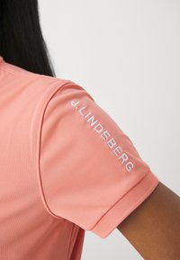 Närbild av en person som bär en korallfärgad kortärmad skjorta med "J. Lindeberg" broderat i vitt på ärmen.