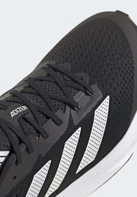 Gros plan d'une chaussure de course noire avec des bandes blanches perforées, des lacets noirs et une semelle blanche portant les inscriptions Lightsource et Adizero.