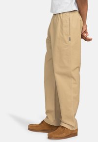 Beige, løst siddende bukser med elastisk talje og sidelomme, lavet af letvægtsstof, parret med brune mokkasiner.