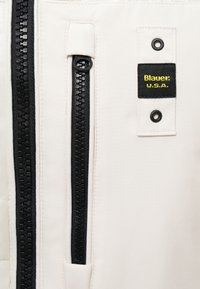 Tissu de veste blanche avec une fermeture éclair noire bien visible et une étiquette qui indique "Blauer U.S.A." avec des détails jaunes. Texture lisse dans l'ensemble.
