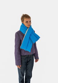 Modrá vystužená šatka s krémovou spodnou stranou. Vlastnosti zahŕňajú mäkkú textúru a nastaviteľné zapínanie. Nosená na fialovej bunde a tmavých džínsoch.